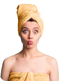 Woman with playful expression, puckering lips, and wide eyes, wrapped in a yellow towel turban and another towel around her body against a plain background, conveying fun, relaxation, and a carefree spirit.