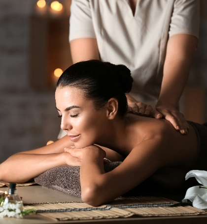 Serene spa scene with a woman receiving a massage, lying peacefully on a table under soft ambient lighting. The hands of the massage therapist working on her back are visible, enhancing the moment of tranquility. Surrounding elements like a candle and natural decor contribute to the calming atmosphere, embodying relaxation, self-care, and wellness.