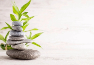 Smooth stones carefully stacked in a serene and balanced arrangement, varying in size and color, against a backdrop of green bamboo leaves and a soft, light wood texture, evoking tranquility and mindfulness, reminiscent of meditation or spa settings.