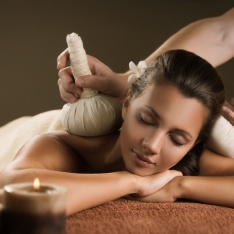 Relaxing spa scene with an individual receiving a massage on a table, covered with a soft cloth, as a practitioner uses a herbal compress on their shoulders and neck. A lit candle casts a warm glow, enhancing the tranquil atmosphere and embodying the serenity of therapeutic massage.