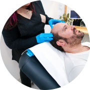 Man receiving a scalp treatment, featuring short hair and a light beard, looking relaxed as he reclines slightly. A professional wearing blue gloves is gently massaging or applying a treatment to his scalp with their fingers. The scene is set in a modern salon or spa environment, characterized by a clean background and a comfortable treatment chair. This demonstrates a soothing scalp care session, highlighting a focus on relaxation and wellness.