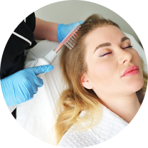 Woman relaxing during a hair treatment session, featuring long, light blonde hair and closed eyes, suggesting enjoyment of the process. A person with blue gloves uses a specialized tool with a comb-like applicator to treat her scalp or hair. She is lying back on a treatment table, covered with a white towel or robe, creating a spa-like atmosphere. Overall, the scene reflects a serene hair care experience focused on relaxation and rejuvenation.