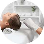 Woman lying back in a salon undergoing a hair treatment or coloring process, appearing relaxed. A person in white gloves applies a product to her hair using a brush or applicator. The background features a clean, modern salon with light-colored decor and neatly arranged items, creating a soothing atmosphere. Overall, the scene conveys a professional hair care experience focused on relaxation and treatment.
