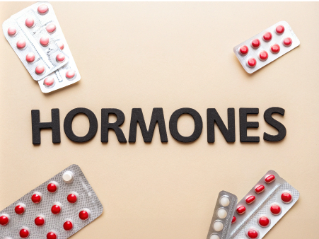 The word 'HORMONES' prominently displayed in bold black letters against a soft beige background, surrounded by several blister packs of medication. The top left and top right packs contain red and white pills, while a larger pack at the bottom left features a mix of red pills, and another at the bottom right has red and white pills. This arrangement creates a visual focus on the term 'HORMONES,' suggesting a theme related to health, wellness, or pharmacology. The overall aesthetic is clean and organized, evoking a clinical yet engaging feel.
