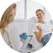 Friendly interaction between a healthcare provider and a patient in a clinical setting. The healthcare provider, a woman with long blonde hair wearing a white blouse, is seated facing the patient, holding a clipboard and listening attentively while wearing blue medical gloves. The patient, also a woman, is comfortably seated in a treatment chair, dressed in a casual white t-shirt and light-colored jeans. She is smiling and gesturing with her hand, indicating active participation in the conversation. The bright and airy background, with natural light filtering through large windows, creates a welcoming atmosphere. The overall scene conveys warmth, professionalism, and open communication in a healthcare environment.