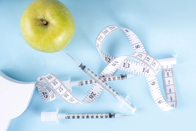 Composition centered around health and wellness themes featuring a bright green apple in the top left corner, symbolizing nutrition and healthy eating. A flexible measuring tape is coiled around, with visible measurements in both inches and centimeters, indicating a focus on monitoring weight or body metrics. Three syringes are prominently displayed in the middle, suggesting medical or therapeutic applications, possibly related to insulin or other health-related injections. The soft blue background provides a calm and clinical atmosphere, contrasting well with the bright apple and the white syringes and measuring tape. The overall arrangement conveys themes of health management, emphasizing the balance between nutrition and medical care.