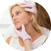 Beauty or skincare consultation in a clinical setting featuring a young woman with long, wavy blond hair seated with a thoughtful expression, looking slightly upward. She wears a white top, contributing to the clean and fresh aesthetic of the scene. A hand wearing a pink glove gently touches the patient's jawline, indicating an assessment of facial features or a discussion about treatment. The soft, blurred background keeps the focus on the woman and the interaction, conveying a sense of care and professionalism, with an emphasis on beauty treatments or skin health.