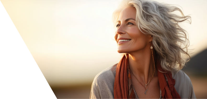 Smiling woman with beautiful silver hair, conveying joy and contentment. She has a serene expression, gazing slightly upwards as if enjoying a pleasant moment or contemplating something uplifting. Dressed in a light-colored, flowing top and a warm-toned scarf draped around her neck, she radiates a relaxed and natural appearance. The softly blurred background features a warm glow, likely from a sunset, enhancing the warmth and positivity of the scene. Overall, the image emphasizes happiness and tranquility, celebrating beauty and confidence at any age.