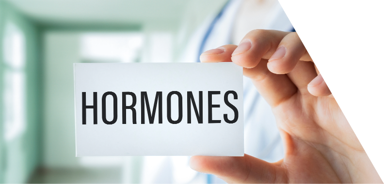 Close-up of a healthcare professional holding a card that prominently displays the word 'HORMONES.' The card is white with bold, black text that contrasts sharply, making the word 'HORMONES' easily noticeable. A partially visible hand holds the card, while the background features a soft-focus clinical setting, likely a medical office or clinic, with green walls and bright lighting that suggest a clean and professional environment. Overall, the image conveys a clinical focus on hormones, possibly indicating a discussion about health, wellness, or endocrinology in a supportive healthcare context.
