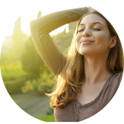 Serene outdoor scene featuring a young woman enjoying nature. She has long, wavy hair and stands with her eyes closed, wearing a peaceful smile. Her arms are raised behind her head, suggesting a relaxed and carefree moment. Warm, golden sunlight creates a soft glow around her, enhancing the tranquility of the scene. The background includes lush greenery with blurred trees and foliage, evoking a calm, natural environment. Overall, the image captures a moment of joy and relaxation, highlighting a connection with nature and well-being.