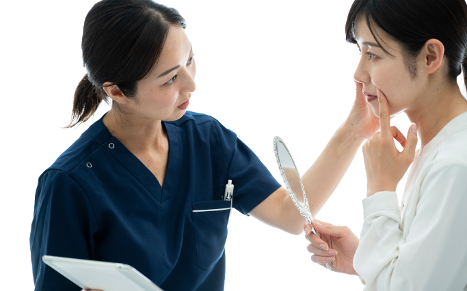 Professional interaction between a healthcare provider and a patient in a clinical setting. The healthcare provider, a woman dressed in a dark blue scrubs top, has dark hair pulled back and is focused on the patient, demonstrating care. She holds a tablet in her left hand, suggesting she is reviewing information or discussing treatment options. The patient, a young woman in a light-colored blouse, is examining her reflection in a handheld mirror, appearing engaged and thoughtful as she touches her face, indicating a discussion about cosmetic or skincare concerns. The neutral background allows focus on their interaction, conveying professionalism and trust in a healthcare environment, suggesting a consultation aimed at enhancing the patient's well-being.