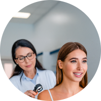 Healthcare professional wearing glasses and a light blue shirt positioned behind a young woman in a clinical setting. The professional appears focused while using a device, likely for a skin examination. In the foreground, the young woman smiles and looks relaxed, with long, wavy hair, suggesting a positive experience. The softly blurred background directs attention to their interaction, creating an overall atmosphere of professionalism and care in a medical or cosmetic context.