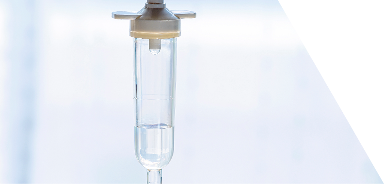 Intravenous (IV) drip bag prominently displayed in a clinical setting. The translucent bag allows a clear view of the sterile liquid inside, which appears to be saline or a nutrient solution. At the top, a white connector facilitates attachment to an IV line, while the bottom features a funnel-like opening for liquid flow. The softly blurred background emphasizes the IV bag, with light filtering in to create a clean and sterile atmosphere. Overall, the image conveys a sense of medical care, highlighting the importance of IV therapy in healthcare settings.