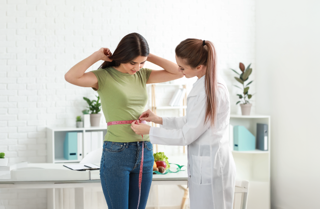 Nutritionist measuring a woman's waist with a tape in a bright health clinic; white brick wall and plants in the background create a welcoming wellness environment. Table with fresh fruits and vegetables symbolizes healthy lifestyle and diet management.