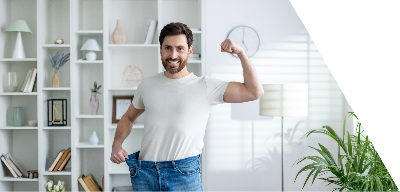 Confident man flexing his bicep while holding oversized jeans, showcasing significant weight loss; dressed in a white t-shirt and jeans in a modern, well-lit living space with plants and decor, emphasizing fitness success and a healthy lifestyle.