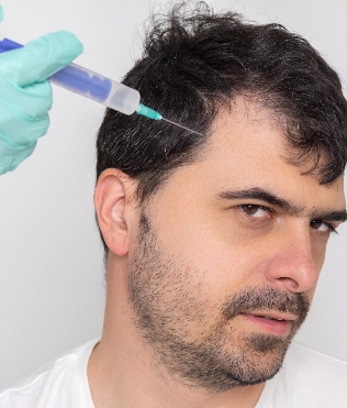 Man with short, dark hair looking skeptical as a syringe, held by a hand in a light blue glove, is poised near his head, suggesting a medical or cosmetic procedure, against a neutral background, conveying themes of hair restoration or cosmetic treatment.