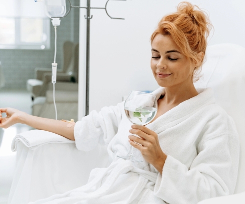 Woman in a white bathrobe sitting comfortably with a relaxed expression, holding a glass of water with lemon or lime, receiving an IV drip treatment, in a tranquil and minimalistic setting, emphasizing wellness, self-care, and relaxation.