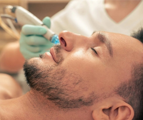 Man undergoing a facial treatment with a skincare device by a practitioner in light blue gloves, in a serene spa or clinic setting, emphasizing themes of self-care and grooming.
