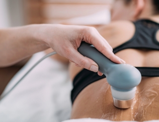 Close-up of a hand holding a skincare device applied to a person's back, who is wearing a black top and lying face down, with gel or oil for smooth movement, focusing on relaxation and body care against a softly blurred background.