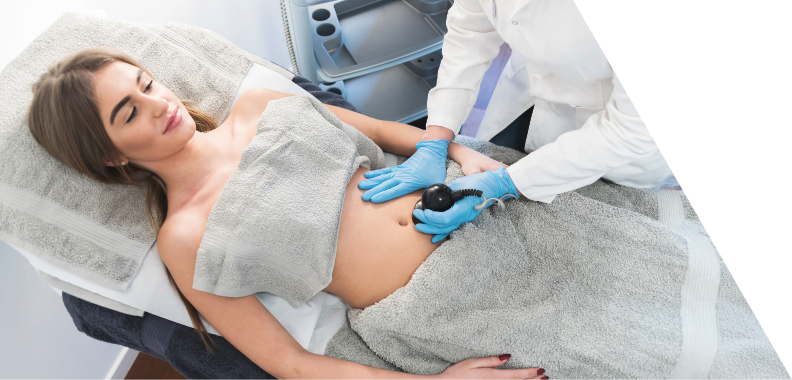 Relaxed woman lying on a treatment bed covered with a gray towel while a professional in a white coat administers an abdominal procedure with a handheld device; clean, well-lit environment conveying professionalism and comfort in a medical setting.