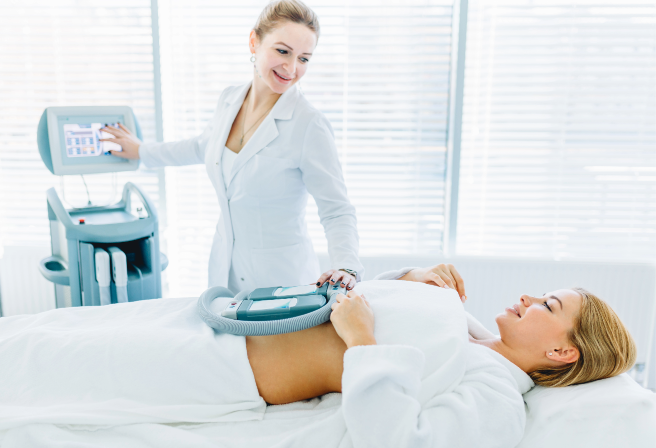 Clinical setting with a relaxed woman on a treatment bed, covered in a white blanket, smiling slightly during a procedure; a professional in a white lab coat uses a handheld device on her abdomen in a bright, clean environment with natural light, emphasizing care and professionalism.