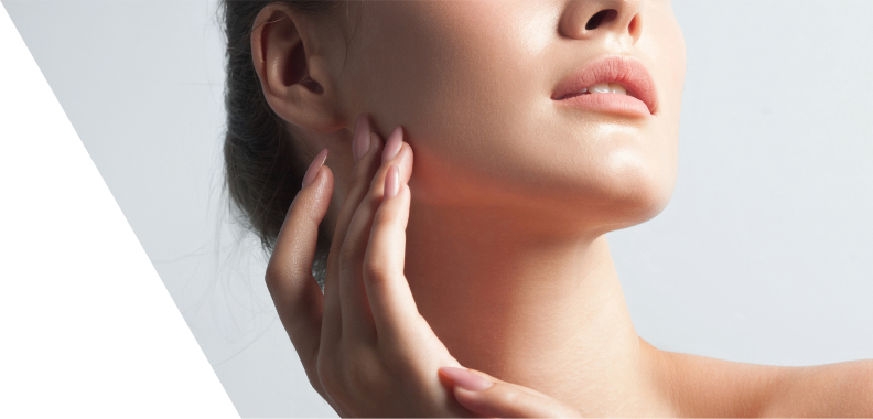 Close-up of a person's face, focusing on the jawline and neck area. The subject, likely a woman, has a serene expression with soft features and a natural, radiant complexion. Her hand gently touches her jaw, conveying elegance and contemplation. The minimal background emphasizes the soft lighting that highlights the contours of her face. The overall aesthetic is polished and sophisticated, suggesting themes of beauty and self-care